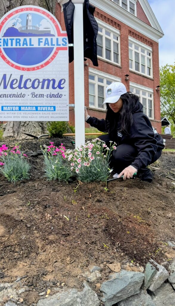 cepeda1 | The Diamant Foundation Person kneeling to plant pink and white flowers in a garden bed beside a 'Welcome Central Falls' sign in front of a brick building.