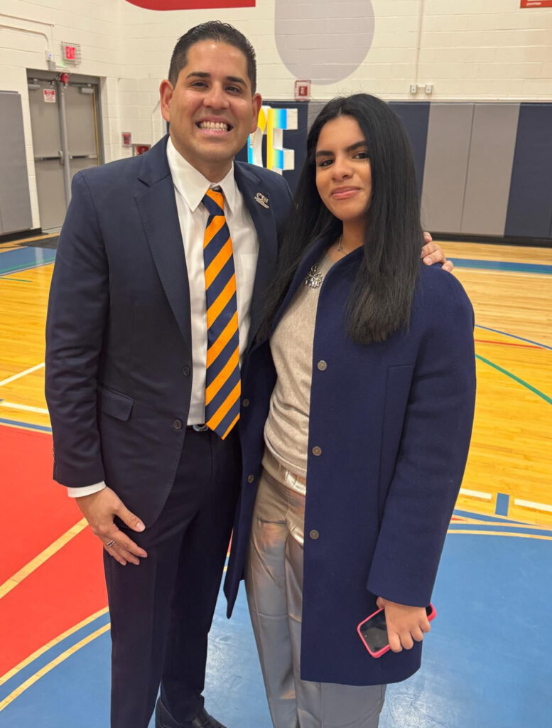 cepeda3 | The Diamant Foundation Two people stand together in a gym, smiling for the photo; man in a navy suit with an orange-and-navy striped tie, woman in a dark coat holding a pink phone.